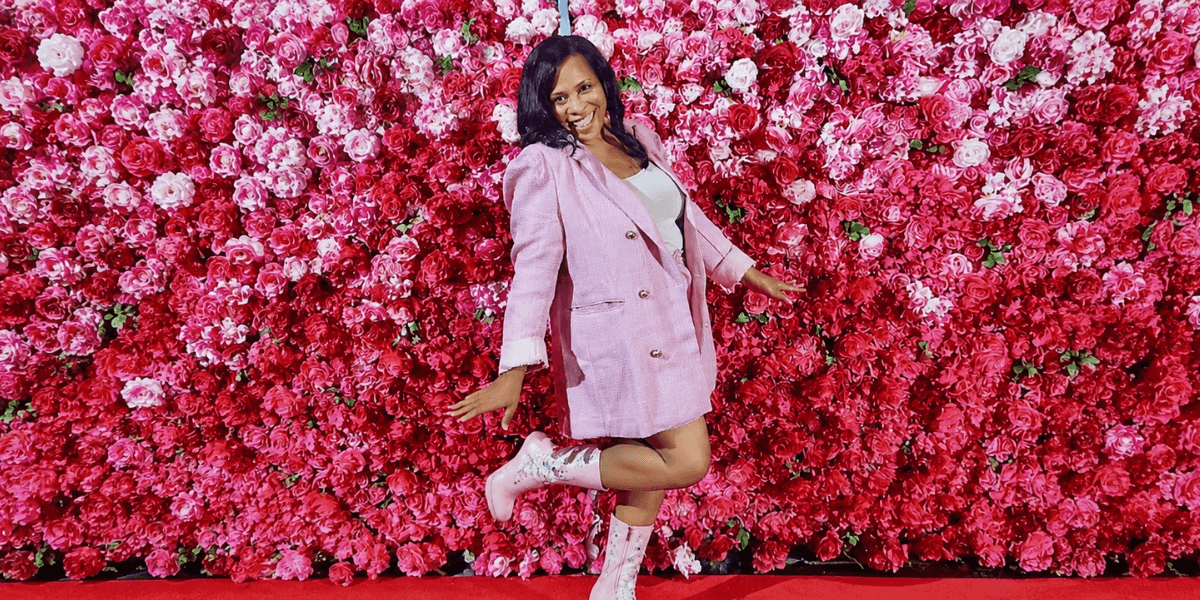 Katrina Frazier posing in a pink blazer and floral boots against a wall of pink flowers.