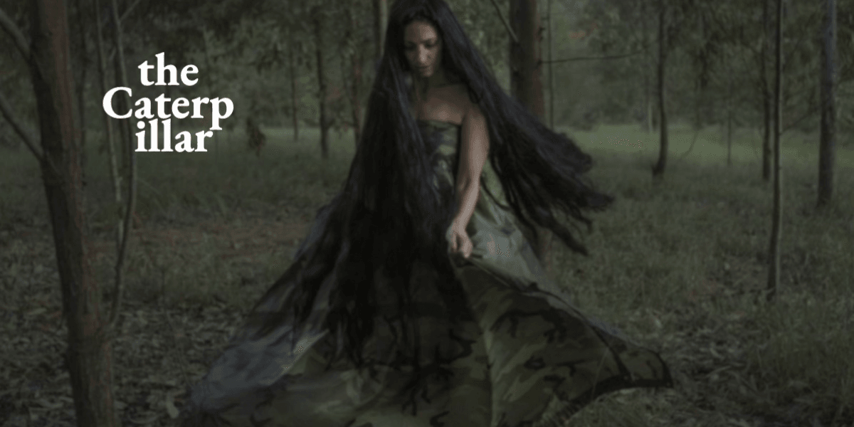 A woman in a camouflage dress stands in a forest with long hair flowing.