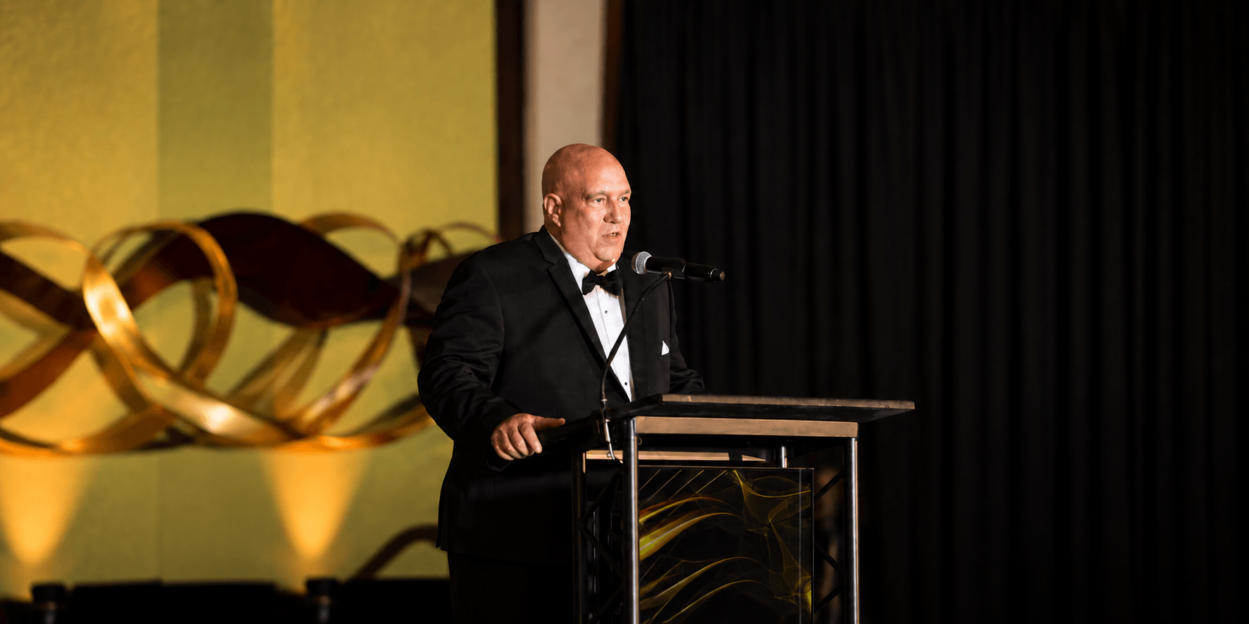 A man in a tuxedo speaks at a podium during an event.