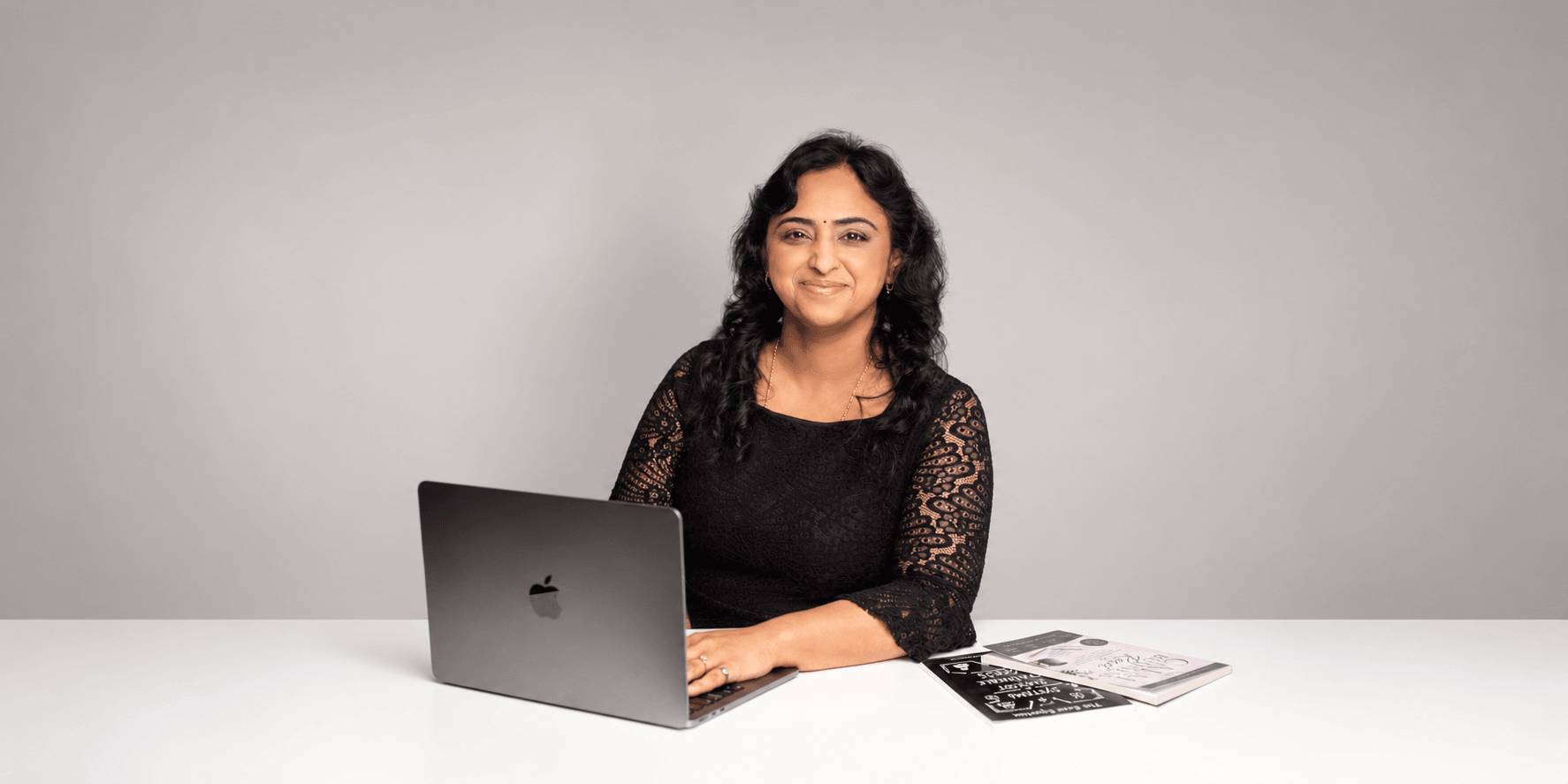A woman with black curly hair sits at a desk with a laptop and notebooks