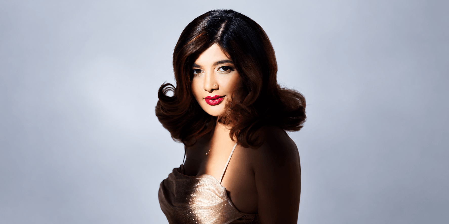 Woman with wavy hair in a gold dress, softly lit against a light background