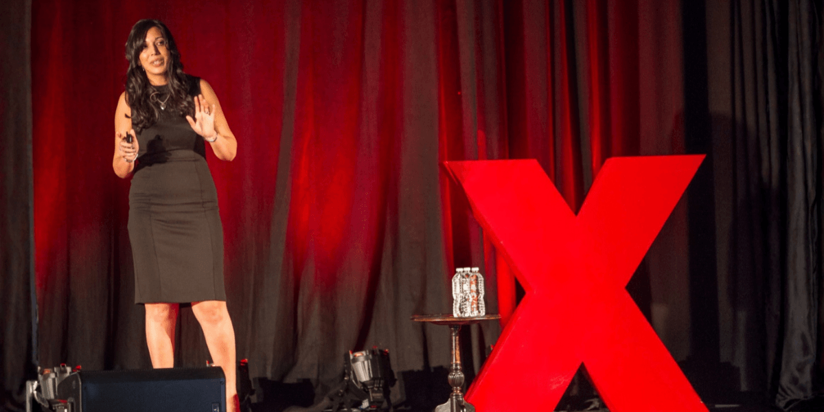 Woman speaking on stage beside a large red TEDx logo and water bottles
