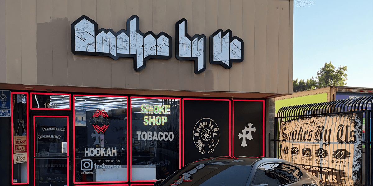 Storefront of a smoke shop with "Smokes By Us" sign and glass windows.